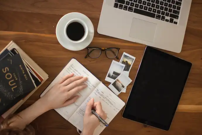 overhead view of coffee cup, laptop, hands writing in notebook